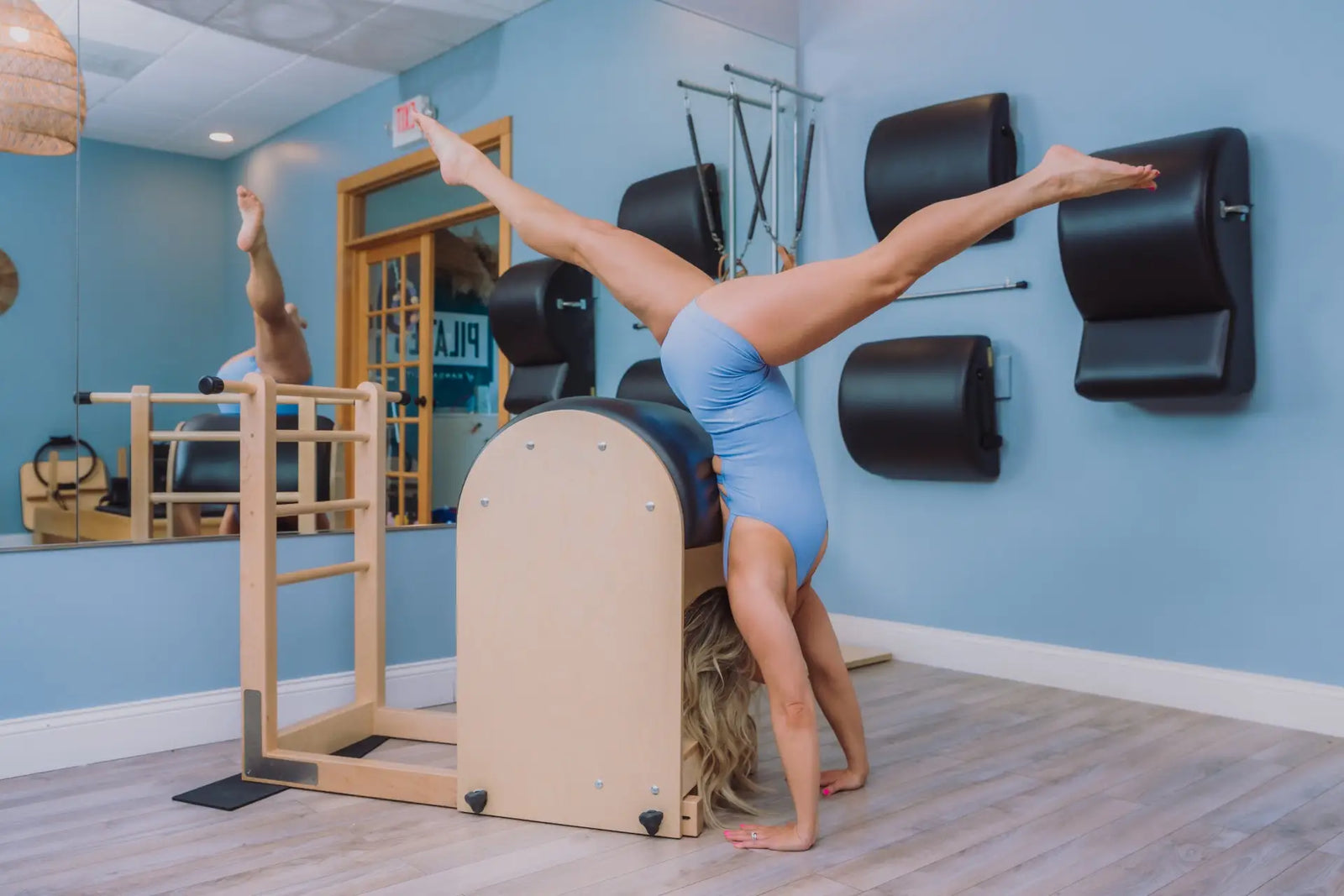 Person in blue leotard performing a split inversion over a wooden Pilates Ladder Barrel in a studio.