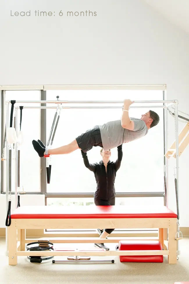 A man exercises on a Pilates Cadillac with Alexandra Adams assisting in a bright studio. Text reads: "Lead time: 6 months."